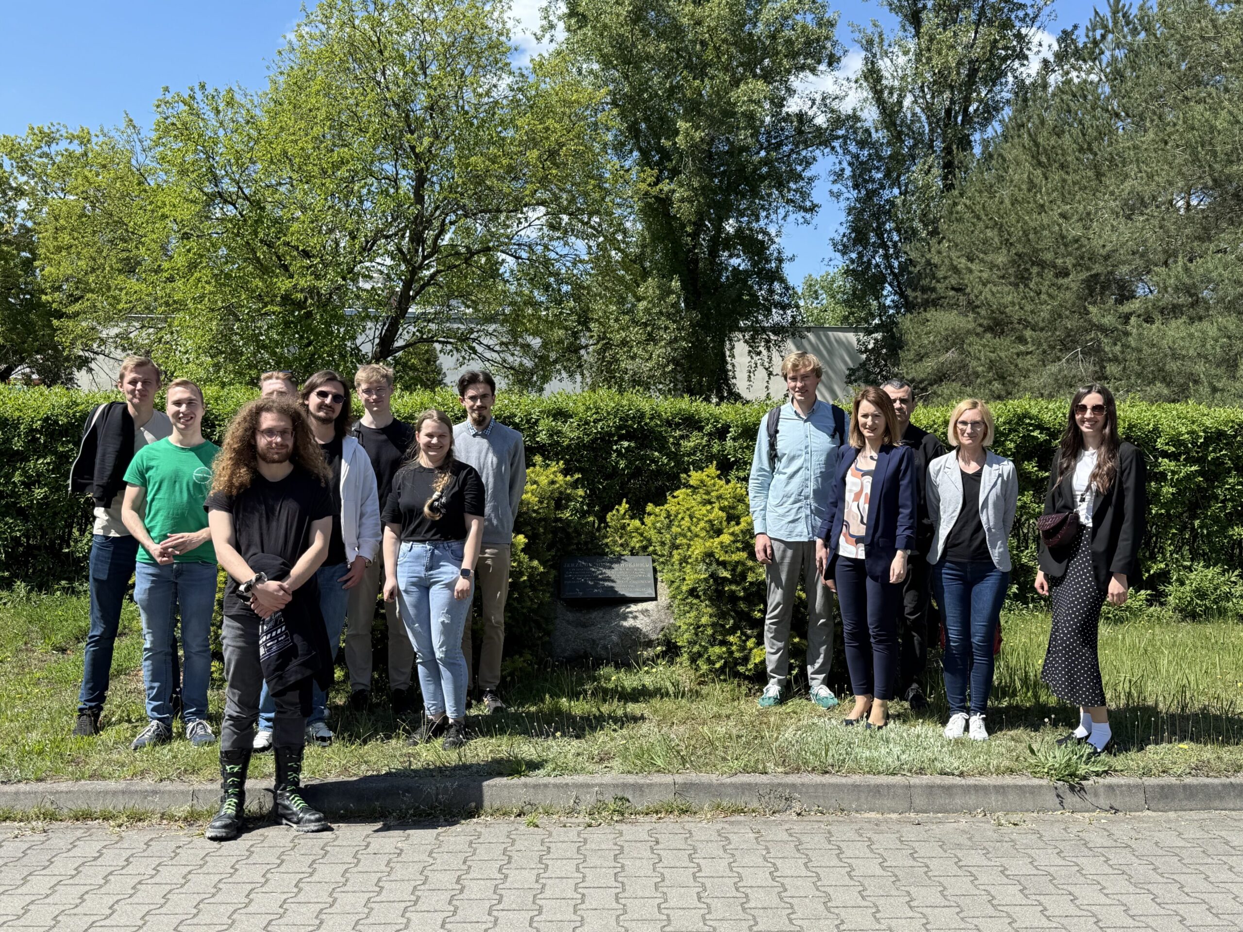 Grupa studentów i pracowników AGH pozuje do wspólnego zdjęcia plenerowego w słoneczny dzień. W tle widać zielone drzewa i żywopłot, co nadaje fotografii naturalny charakter. Wszyscy uczestnicy uśmiechają się, co świadczy o przyjaznej atmosferze spotkania.