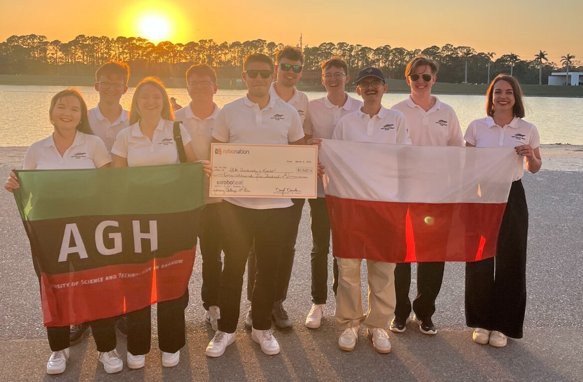 Grupa studentów w białych koszulkach polo z logo drużyny, stojących na tle zachodzącego słońca nad wodą. Trzymają flagi AGH i Polski oraz symboliczny czek z zawodów RoboBoat.
