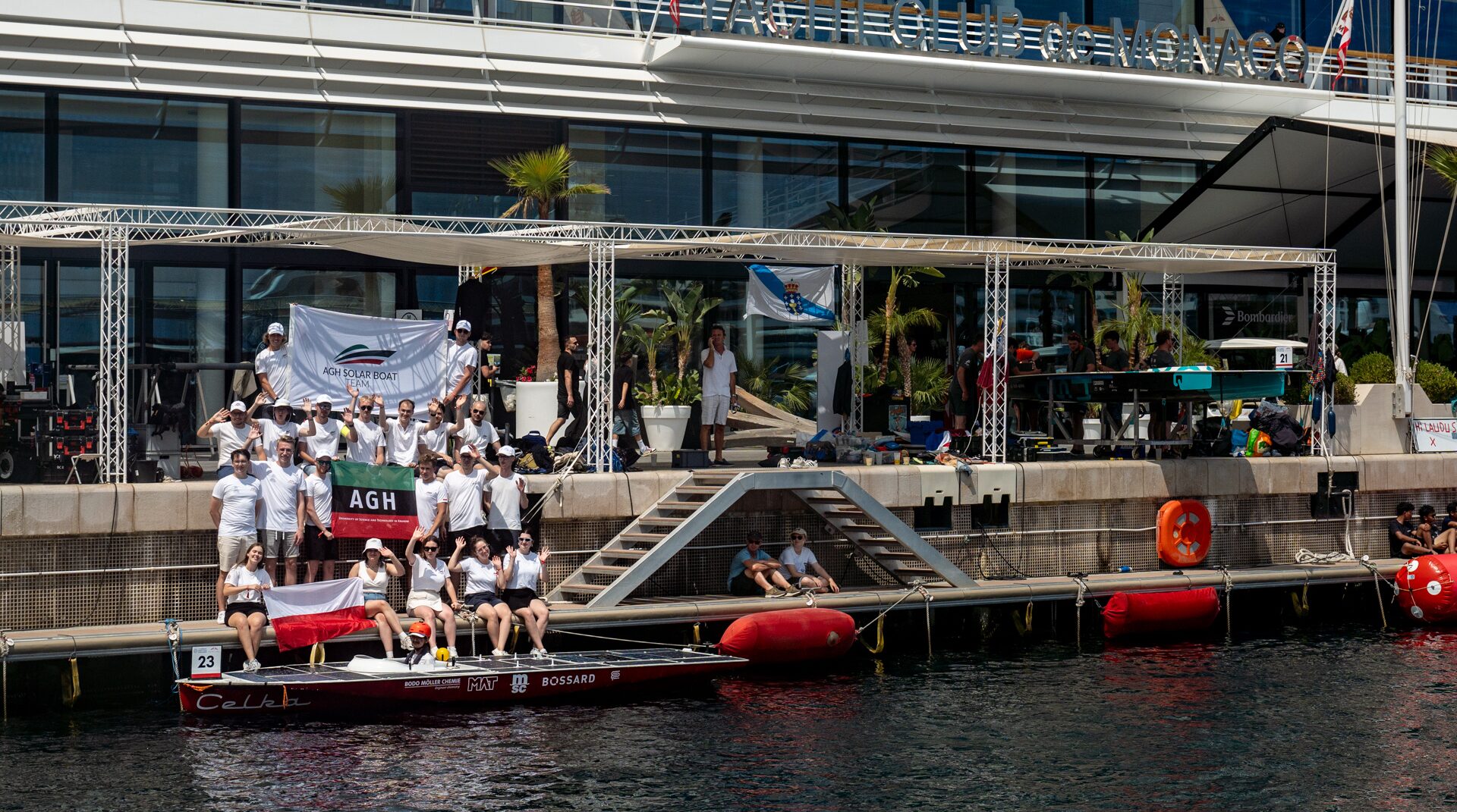 Grupa ludzi w białych koszulkach z flagami "AGH SOLAR BOAT TEAM" pozuje na pomoście obok czerwono-białej łodzi solarnej "Celka" z numerem 23.