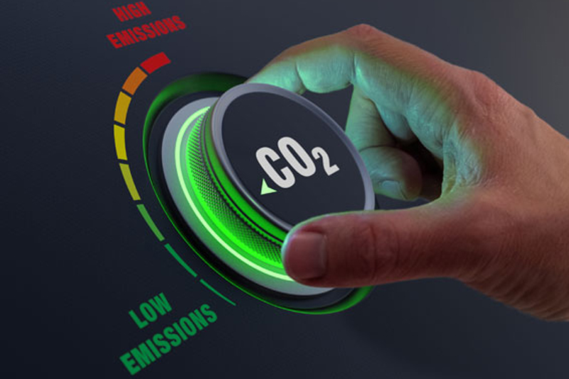 Ręka obracająca pokrętło oznaczone jako CO₂ w kierunku zmniejszenia emisji. Pokrętło otoczone jest kolorową skalą od czerwonej (wysoka emisja) do zielonej (niska emisja), na tle zielonych, rozmytych okręgów